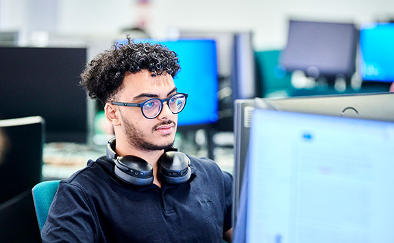 Ahmed Mostafa, a Computer Science student specializing in Game Engineering, sits working at a computer, in one of our computer science labs. Ahmed has a light-medium skin tone, black curly hair, and wears glasses. He